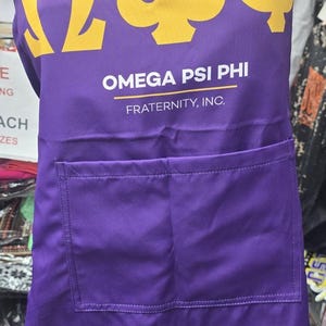 May include: A purple apron featuring a yellow Omega Psi Phi logo and text. The apron has a front pocket and the words "OMEGA PSI PHI FRATERNITY, INC." are printed below the logo.