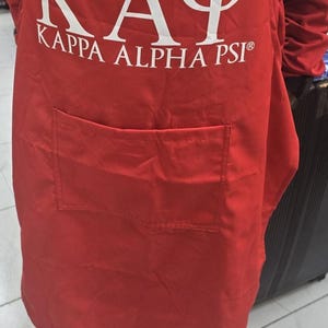 May include: Red apron with a large white "KAY" and "KAPPA ALPHA PSI" text. The apron has a front pocket and is worn by a person. The apron is made of a smooth fabric.