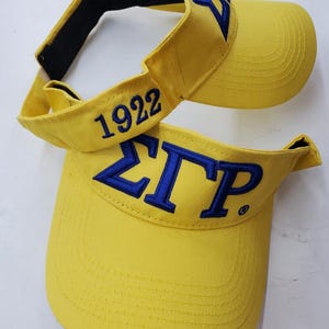 May include: Two yellow visors with blue embroidered Greek letters and the number 1922. The visors have a curved brim and an adjustable black strap. The text "ΣΓΡ" is embroidered on the front of one visor.