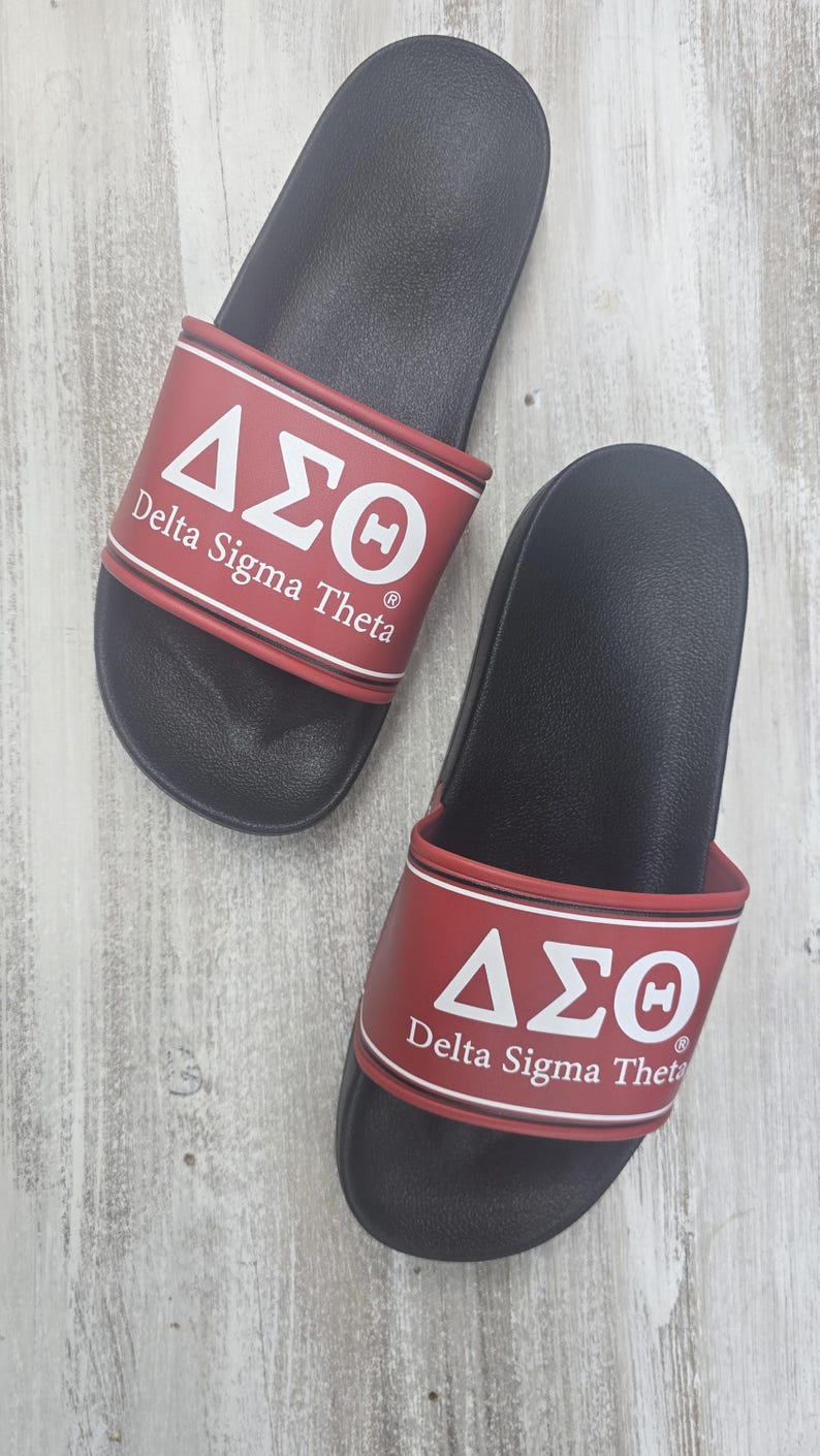 May include: Pair of black slide sandals with red straps. The straps feature the Greek letters Delta, Sigma, and Theta in white, along with the words "Delta Sigma Theta". The sandals are on a light wood-grain surface.