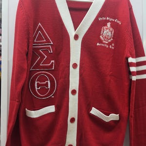 May include: A red V-neck cardigan with white trim, featuring the Greek letters Delta Sigma Theta embroidered on the right side and the Delta Sigma Theta Sorority, Inc. crest on the left chest. It has five red buttons and two front pockets.