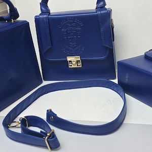 May include: Three structured royal blue handbags with a smooth finish, gold-tone metal hardware, and top handles. Two bags feature a front flap with a gold clasp and an embossed crest logo. A detachable, adjustable shoulder strap lies in the foreground.