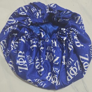 May include: A royal blue satin bonnet featuring a repeating white print of the Greek letters Z&Phi;B and the year 1920. The hair accessory has a gathered, elasticized edge and a smooth, shiny finish throughout the fabric surface.