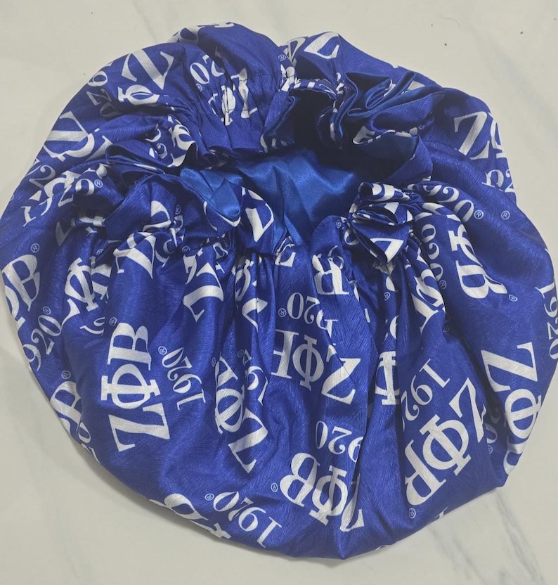 May include: A royal blue satin bonnet featuring a repeating white print of the Greek letters Z&Phi;B and the year 1920. The hair accessory has a gathered, elasticized edge and a smooth, shiny finish throughout the fabric surface.