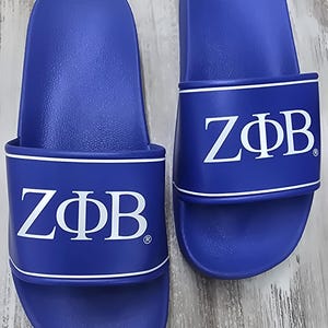 May include: A pair of blue slides with white trim and the letters ZΦB in white. The slides are made of a smooth material and are designed for casual wear. The footwear is displayed on a light-colored surface.