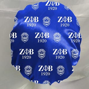 May include: A royal blue satin bonnet featuring a repeating white print of the Greek letters Z&Phi;B, the year 1920, and a crest emblem. The fabric has a smooth, shiny finish and a gathered elastic edge for a secure fit around the head.