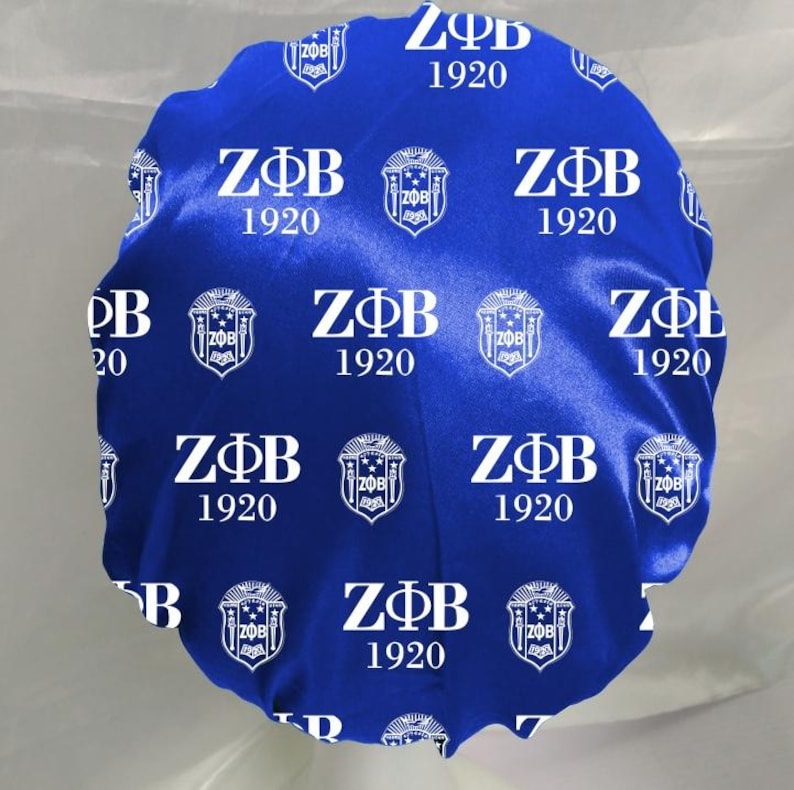 May include: A royal blue satin bonnet featuring a repeating white print of the Greek letters Z&Phi;B, the year 1920, and a crest emblem. The fabric has a smooth, shiny finish and a gathered elastic edge for a secure fit around the head.