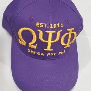 May include: A purple baseball cap with gold embroidered text. The text includes the Greek letters Omega, Psi, and Phi, along with "EST. 1911" and "OMEGA PSI PHI". The cap has a curved brim and a button on top.