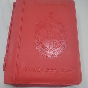May include: A red leather binder with the text "Delta Sigma Theta" and "Sorority, Incorporated" embossed on the cover. The binder features a zipper closure and a crest emblem. The binder is a vibrant shade of red.