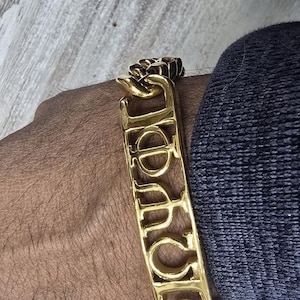 May include: A gold-toned metal chain bracelet featuring a central rectangular plaque with cut-out Greek letters Phi, Phi, and Omega. The bracelet is constructed with thick, interlocking curb links and has a polished, reflective finish throughout.