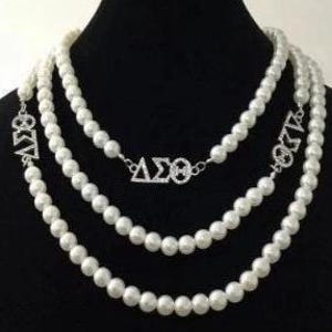 May include: Three-strand pearl necklace with silver-tone Greek letter pendants. The necklaces are draped on a black mannequin. Each strand is composed of white, round pearls. The pendants feature the Greek letters Delta, Sigma, and Omega.