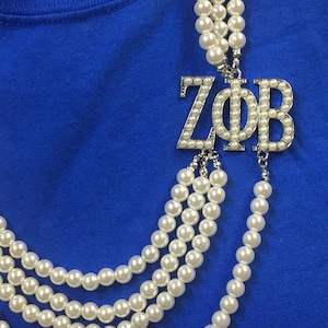 Puede incluir: Un collar de perlas de múltiples hilos con un colgante plateado. El colgante presenta las letras griegas Z, Φ y B, adornadas con pequeñas perlas. El collar se muestra sobre un fondo azul.