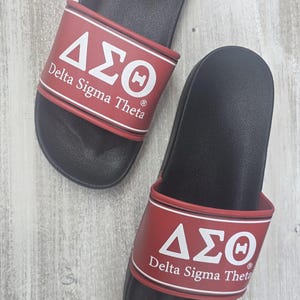 May include: Pair of black slide sandals with red straps. The straps feature the Greek letters Delta, Sigma, and Theta in white, along with the words "Delta Sigma Theta". The sandals are on a light wood-grain surface.