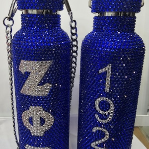 Può includere: Due borracce blu cobalto incrostate di strass. Una bottiglia ha le lettere ZΦB in strass argentati, l'altra ha i numeri 1920. Entrambe le bottiglie hanno cinturini a catena e coperchi argentati.