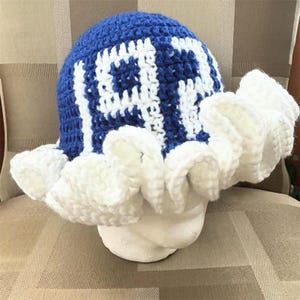 May include: A crochet bucket hat featuring a blue and white geometric square pattern on the crown and a wide, ruffled white brim. The hat is constructed with thick yarn stitches, creating a textured appearance throughout the entire piece.