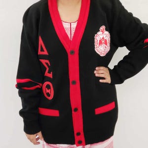 May include: Black cardigan with red trim and a red embroidered Greek letter design on the sleeve. The cardigan has a button closure and two pockets.