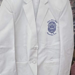 Peut inclure: Un blazer blanc avec un revers cranté, deux boutons et une poche poitrine. Le blazer présente un écusson bleu sur la poitrine avec l'inscription "Zeta Phi Beta Sorority, Inc."