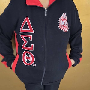 May include: Black fleece jacket with red accents and a zip closure. The jacket features a large embroidered Greek letter design on the front, with the letters Delta, Sigma, Theta, and the letters "AETA ZITHA" below the Greek letters.