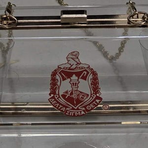 May include: A clear acrylic clutch bag with a gold chain strap and a gold clasp. The bag has a red and white Delta Sigma Theta sorority crest printed on the front.