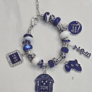 Puede incluir: Un brazalete de plata con cuentas azules y blancas y dijes. Los dijes incluyen un escudo con las letras "ZOB" y el año "1920", un dije cuadrado con las letras "ZOB", un dije con las letras "Z & B" y un dije con las palabras "Desde 1920".