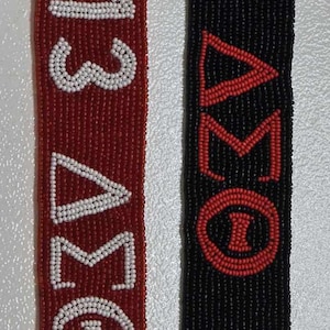 May include: Two beaded straps with the words "OMEGA 1915" in white beads on a red strap and "OMEGA 1915" in red beads on a black strap.