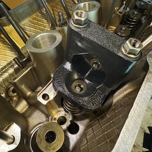 May include: Close-up of a black metal engine component with a silver metal spring and a silver metal nut. The component is mounted on a gold metal engine block.