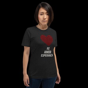 Puede incluir: Camiseta gris oscuro con un corazón rojo texturizado. Las palabras "FE AMOR ESPERANZA" están impresas en blanco debajo del corazón. La camiseta es de manga corta y tiene cuello redondo.