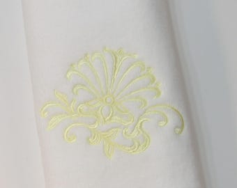 May include: A white hand towel featuring a pale yellow embroidered floral motif in the center. The towel has a soft, plush texture and a decorative fringe along the bottom edge, providing a clean and simple look for any bathroom or kitchen space.