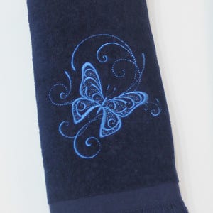 May include: A navy blue hand towel with a light blue embroidered butterfly design. The towel has a fringed edge and is made of a soft, absorbent material. The butterfly is surrounded by swirling accents.