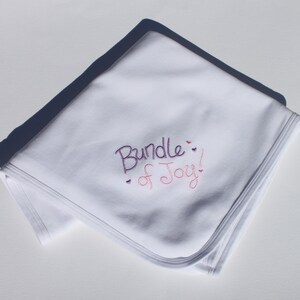 May include: White baby blanket with the embroidered phrase "Bundle of Joy!" in purple and pink script. The blanket is folded, with a shadow cast across the top. The blanket has a stitched edge.