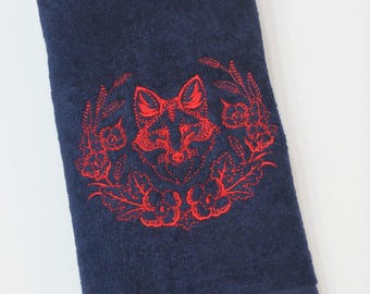 May include: A navy blue rectangular towel featuring a vibrant red embroidered design of a fox head centered within a circular wreath of flowers and leaves. The towel has a textured terry cloth surface and a fringed edge along the bottom hem.
