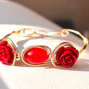 May include: A gold-toned wire bracelet featuring three red accents. Two are rose-shaped, and the center is an oval bead. The bracelet is designed with a delicate, open wirework style, suitable for everyday wear.