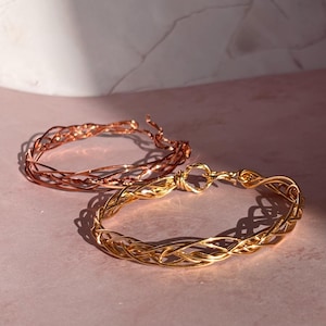 May include: Two wire-wrapped bracelets. One is a copper color, and the other is gold. The bracelets are made of thin, interwoven wires, creating an open, textured design. The gold bracelet has a clasp closure. The bracelets are displayed on a pink surface.
