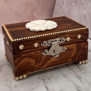 May include: A small, rectangular wooden box with a dark brown finish. The lid features a white flower-shaped embellishment and is trimmed with small gold-colored studs. The box has a silver clasp and is decorated with additional gold studs.