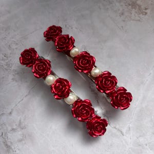 May include: Two hair barrettes with red rose accents and faux pearls. The barrettes are silver-toned and feature multiple red roses and white pearls. The hair accessories are designed for decorative use.