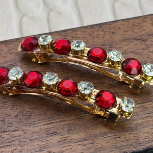 May include: Two gold-toned hair barrettes, each adorned with alternating red and clear crystal-like stones. The barrettes are arranged on a wooden surface, showcasing their decorative design and potential for hair styling.