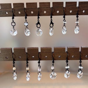 May include: A display of various crystal earrings with black hooks and accents. The earrings feature clear, faceted crystals in different shapes and sizes, some with small pearl or bead accents. The earrings are displayed on a dark brown wooden earring holder.