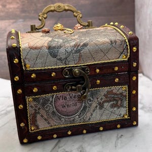 May include: A small, vintage-style wooden chest with a map design on the lid and front. The chest is dark brown with gold-colored accents, including a handle and decorative studs. The text "Via Vee World" is visible on the front.