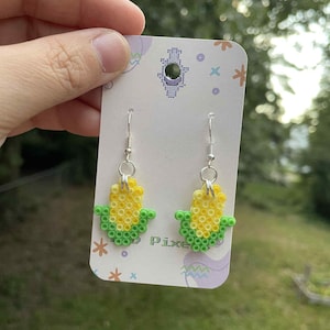 May include: A pair of handmade earrings shaped like corn on the cob. The earrings are made of yellow and green beads, with silver-colored ear wires. The earrings are displayed on a decorative card with the word "Pixel" printed on it.