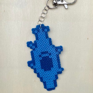 Puede incluir: Un llavero pixelado azul y azul claro con forma de botella. El llavero tiene una cadena de metal plateado y un cierre de metal plateado.