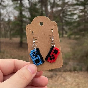 May include: A pair of handmade earrings featuring a video game controller design. One earring is blue with a black design, and the other is red with a black design. The earrings are attached to silver-colored fish hook ear wires.