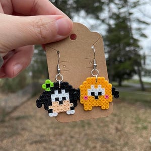 May include: A pair of dangle earrings featuring a black and white perler bead character with a green leaf on its head and a yellow perler bead character with pink cheeks.
