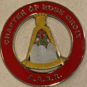 May include: A round, gold-colored emblem with a red border. The text "CHAPTER OF ROSE CROIX" is in gold lettering around the edge. The center features a gold crown, a cross, and an eagle. The letters "A.A.S.R." are at the bottom.