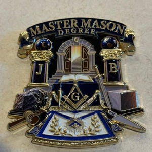 Könnte beinhalten: Ein goldfarbenes Metallabzeichen mit der Aufschrift MASTER MASON DEGREE. Das Design zeigt zwei Säulen mit den Buchstaben J und B, ein offenes Buch, Winkel und Zirkel mit dem Buchstaben G, einen Hammer, ein Schwert, eine Kelle und zwei Steine.