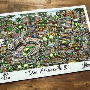 May include: A colorful, detailed illustration of a city, featuring buildings, trees, and a stadium. The artwork is titled "Pubs of Gainesville II" and includes a signature and edition number. The artwork is on a white background.