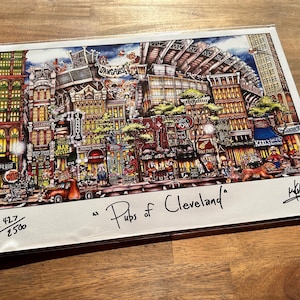 May include: A colorful illustration of a city scene with buildings, streets, and people. The image is titled "Pubs of Cleveland" and is signed by the artist. The image is numbered 427/2500.