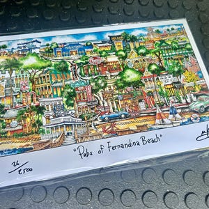 May include: A colorful print titled "Pups of Fernandina Beach" featuring a detailed illustration of a coastal town with buildings, cars, boats, and trees. The artwork is signed and numbered 26/2500.