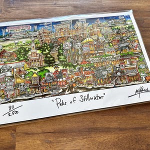 May include: A colorful, detailed art print titled Pubs of Stillwater, featuring a whimsical illustration of various local establishments. The print is numbered 80 out of 2500, includes a signature in the bottom right corner, and is encased in a clear sleeve.