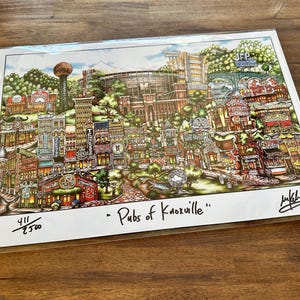 May include: A colorful, detailed art print titled Pubs of Knoxville featuring a dense cityscape illustration of various buildings, storefronts, and a stadium. The print includes a handwritten signature and the edition number 411/2500.