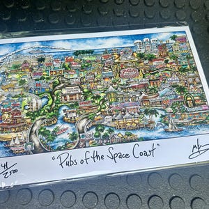 May include: A colorful print titled "Pubs of the Space Coast" depicting a detailed map of a coastal town with various buildings, waterways, and boats. The artwork is signed and numbered 41/2500.